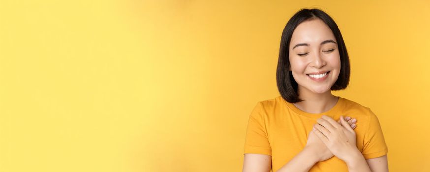 Beautiful asian woman, smiling with tenderness and care, holding hands on heart, standing in tshirt over yellow background.