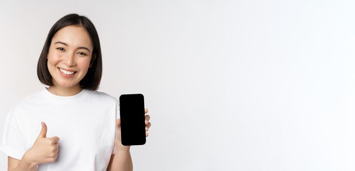 Portrait of beautiful asian woman showing thumb up and smartphone screen, smiling while advertising mobile phone app, white background.
