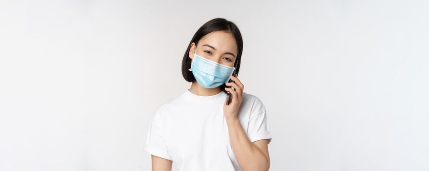 Health and covid-19 concept. Smiling asian girl in medical face mask talking on phone, answer mobile call, standing over white background.