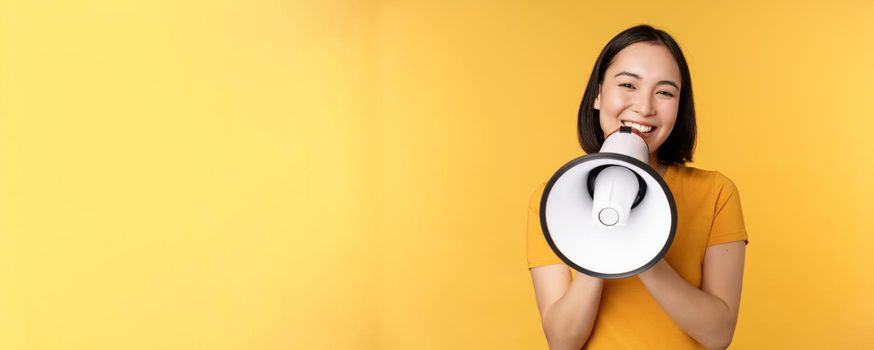 Smiling asian woman standing with megaphone, announcing smth, advertising product, standing over yellow background.