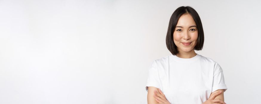 Close up portrait of confident korean girl, student looking at camera with pleased smile, arms crossed on chest, standing over white background.