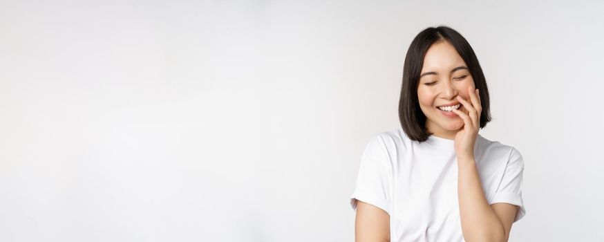 Portrait of young beautiful woman, korean girl laughing and smiling, looking coquettish, standing against white background.