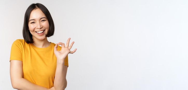 Beautiful young woman showing okay sign, smiling pleased, recommending smth, approve, like product, standing in yellow tshirt over white background.