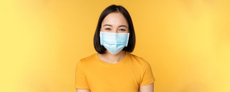 Close up of smiling happy asian woman, wearing medical face mask from covid-19, standing in yellow t-shirt over studio background.