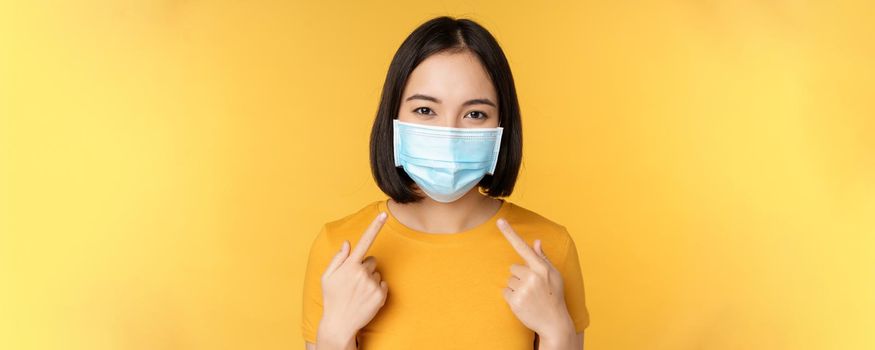 Portrait of smiling asian woman in medical face mask, pointing at her personal protective equipment from covid-19 during pandemic, standing against yellow background.