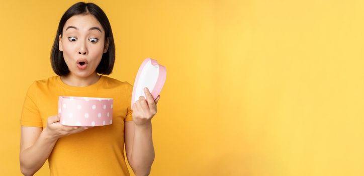 Portrait of excited asian woman, open gift box with surprised happy face, standing over yellow background.