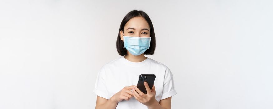 Health and covid-19 concept. Asian girl in medical face mask, using mobile phone application on quarantine, standing over white background.