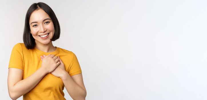 Romantic asian girfriend, holding hands on heart, smiling with care and tenderness, standing in yellow tshirt over white background.