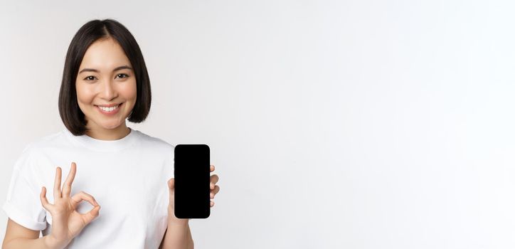 Smiling asian woman showing okay, mobile phone screen, recommending smartphone app, standing over white background.