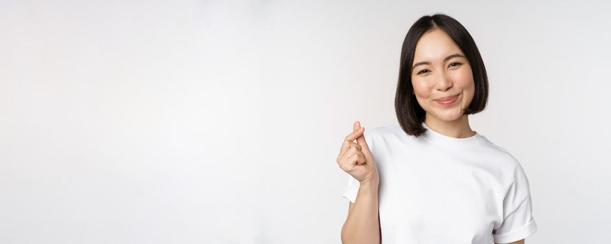 Beautiful asian woman smiling, showing finger hearts gesture, wearing tshirt, standing against white background.