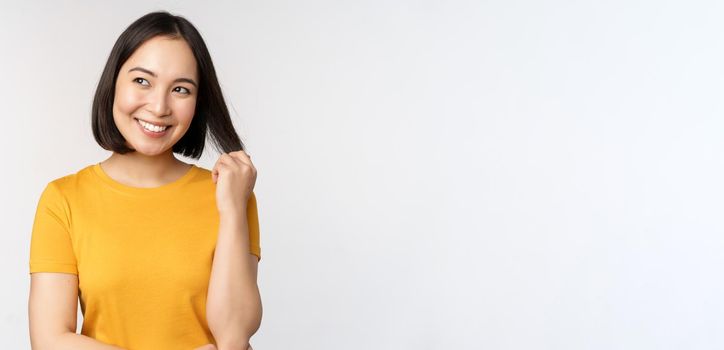 Beautiful romantic asian girl, smiling and playing with hair, looking happy at camera, standing in yellow t-shirt over white background.