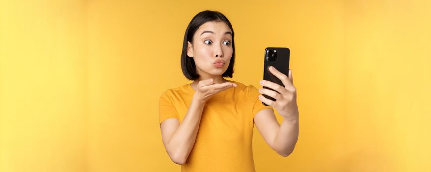 Image of happy, beautiful asian girl video chatting, talking on smartphone application, standing against yellow background. Copy space