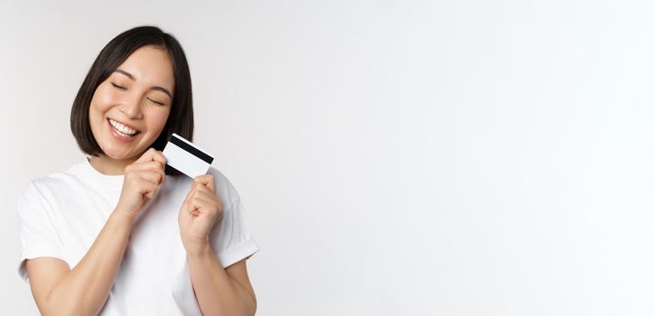 Image of smiling asian woman hugging credit card, buying contactless, standing in white tshirt over white background. Copy space