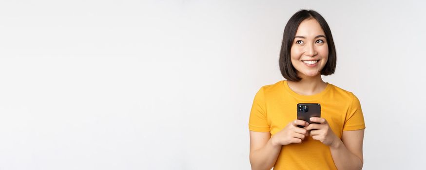 People and technology concept. Smiling asian girl using smartphone, texting on mobile phone, standing against white background.