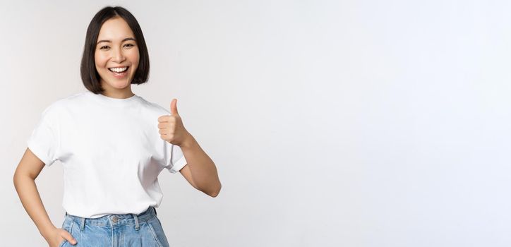 Happy beautiful korean woman, smiling pleased, showing thumbs up in approval, recommending brand or company, standing over white background.