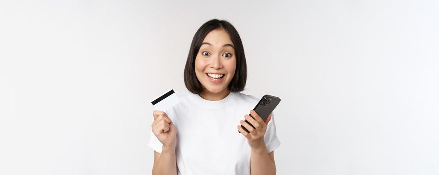 Online shopping. Happy asian woman using credit card and smartphone app, paying on website via mobile phone, white background.