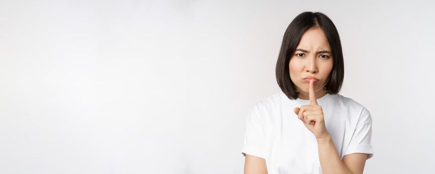 Angry asian girl shushing, keep quiet, taboo silence gesture, press finger to lips and frowning, scolding for being too loud, standing over white background.