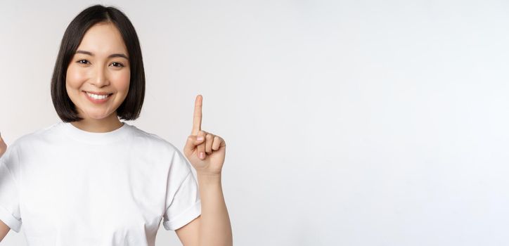 Portrait of enthusiastic young woman, asian girl smiling pointing fingers up, showing advertisement upwards, standing over white background.