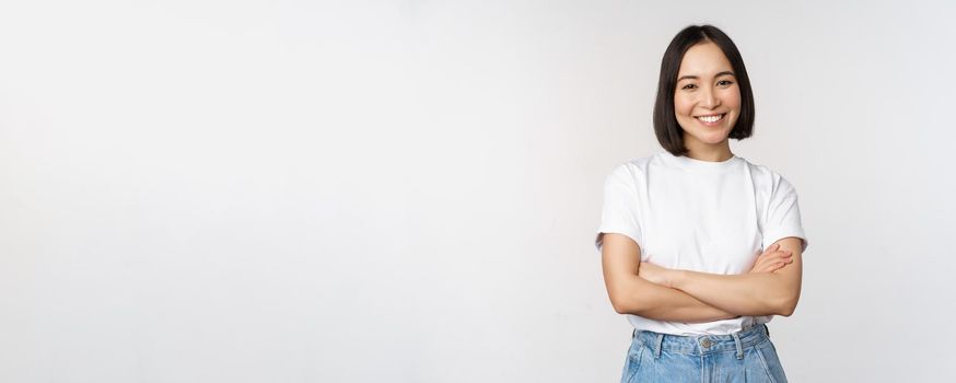 Portrait of happy asian woman smiling, posing confident, cross arms on chest, standing against studio background.