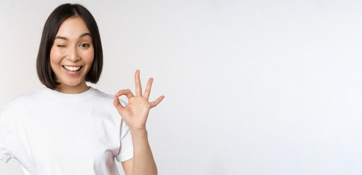 Everything okay. Smiling young asian woman assuring, showing ok sign with satisfied face, standing over white background. Copy space