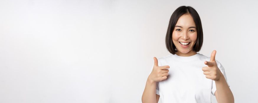 Image of smiling asian girl pointing fingers at camera, choosing, inviting you, congratulating, standing in tshirt over white background.