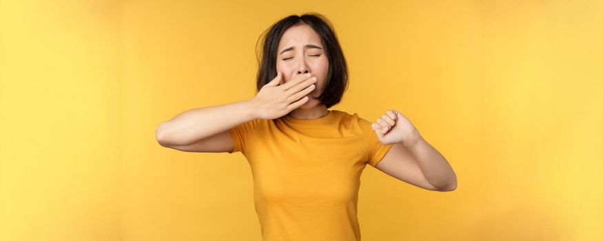 Image of young asian girl student, yawning, feeling tired, standing sleepy agaisnt yellow background. Copy space