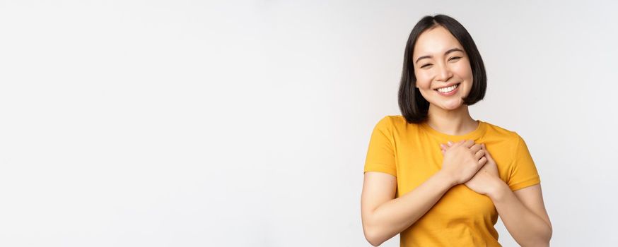 Romantic asian girfriend, holding hands on heart, smiling with care and tenderness, standing in yellow tshirt over white background.