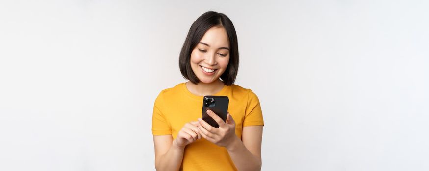People and technology concept. Smiling asian girl using smartphone, texting on mobile phone, standing against white background.