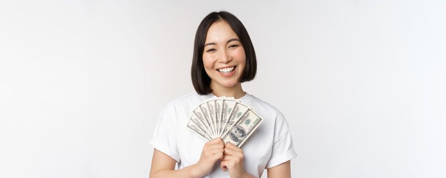 Portrait of smiling asian woman holding dollars money, concept of microcredit, finance and cash, standing over white background.