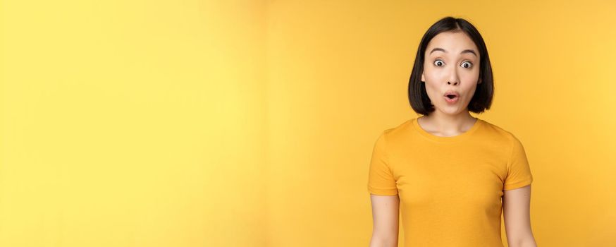 Image of asian girl looking surprised, reacting amazed, raising eyebrows impressed, standing over yellow background.