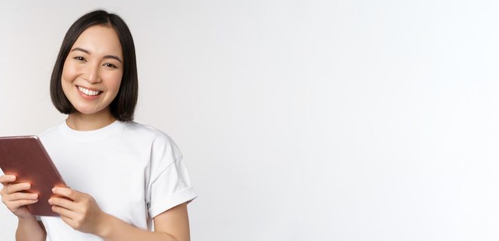 Smiling asian girl with digital tablet, looking happy and laughing, posing in tshirt over white background. Copy space