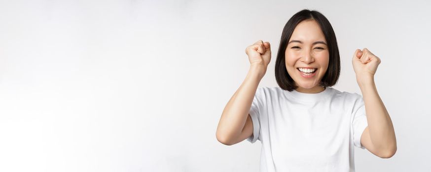 Portrait of enthusiastic asian woman winning, celebrating and triumphing, raising hands up, achieve goal or success, standing over white background.