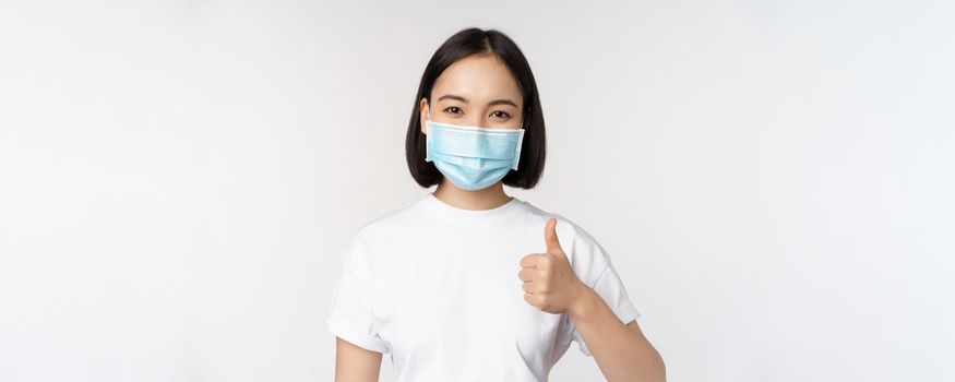 Smiling asian girl in medical mask showing thumbs up, approve something good, praise and compliment company, standing over white background.