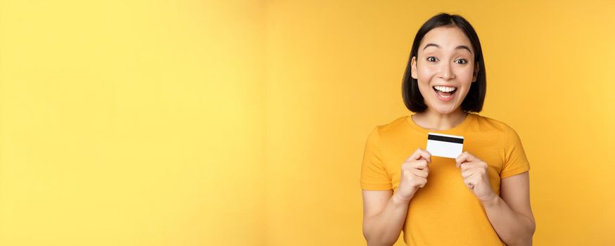 Beautiful asian woman showing credit card and smiling, recommending bank service, standing over yellow background. Copy space