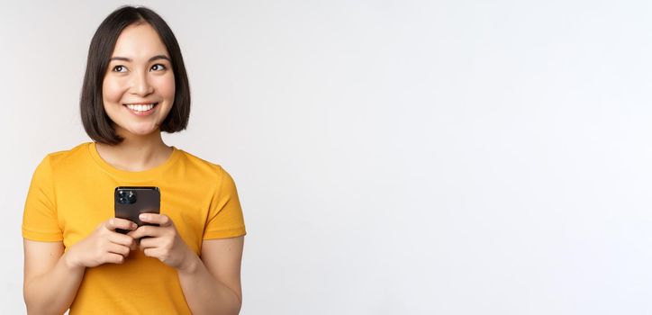 People and technology concept. Smiling asian girl using smartphone, texting on mobile phone, standing against white background.