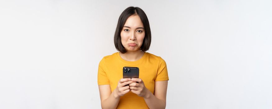 Sad asian woman holding smartphone, looking upset with regret, standing in yellow tshirt against white background.