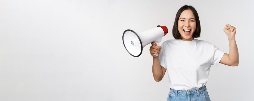 Happy asian woman shouting at megaphone, making announcement, advertising something, standing over white background.