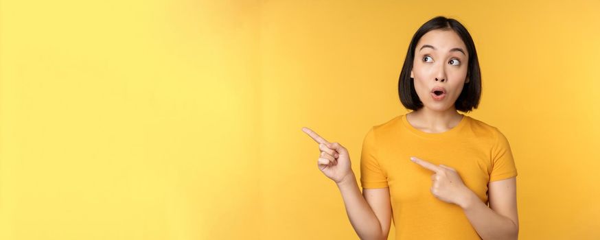Portrait of intrigued asian woman, looking and pointing fingers left at advertisement, showing smth interesting, standing over yellow background.