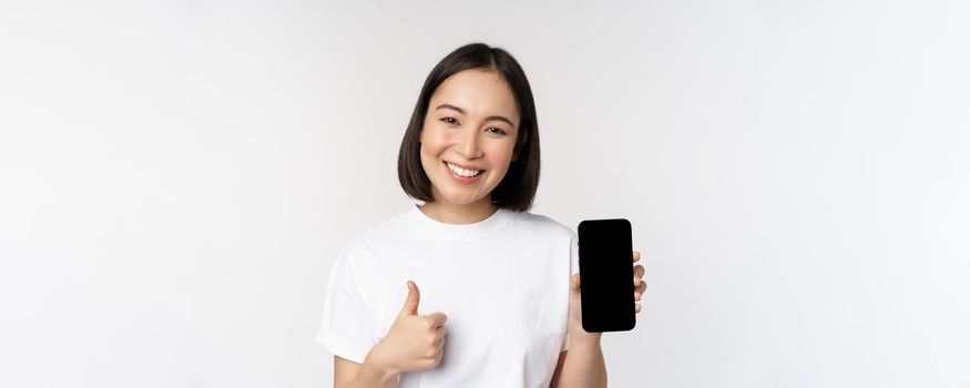 Portrait of beautiful asian woman showing thumb up and smartphone screen, smiling while advertising mobile phone app, white background.