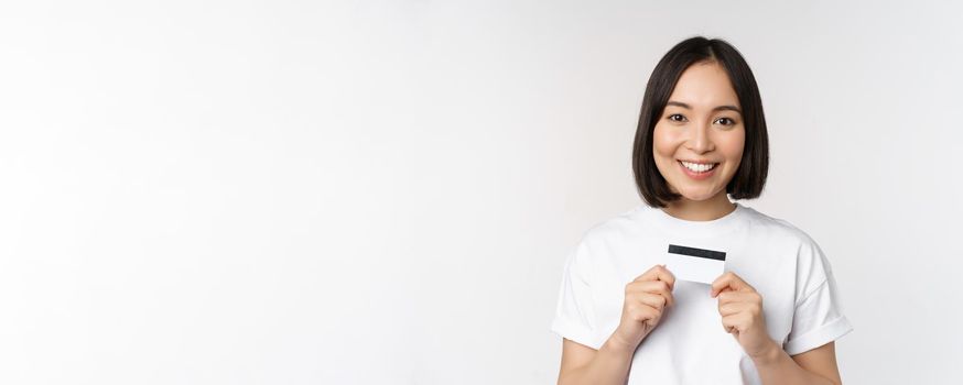 Smiling korean woman showing credit card with happy face, standing in tshirt over white background. Copy space