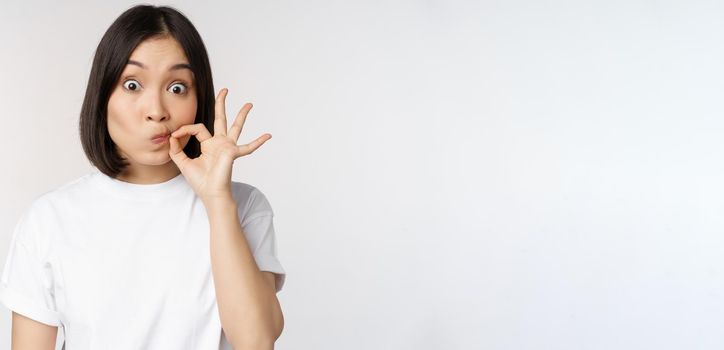Image of young asian woman showing silence gesture, zipping mouth, seal lips, make promise, standing over white background.