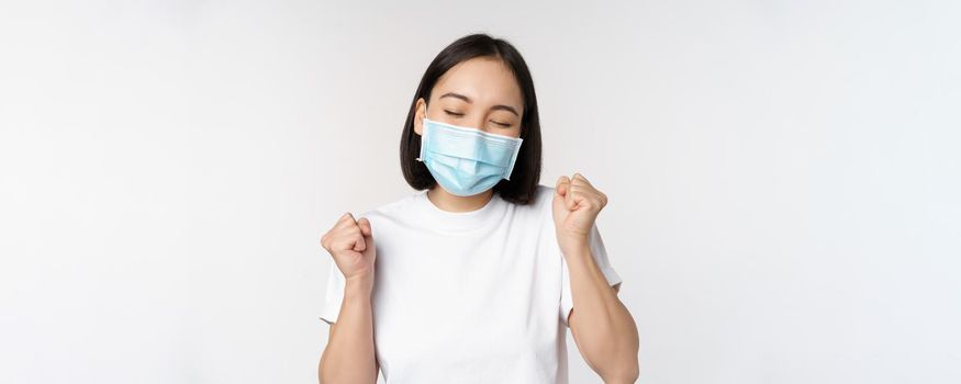 Covid-19, healthcare and medical concept. Enthusiastic asian woman in medical face mask, dancing and celebrating, winning, achieve goal, standing over white background.