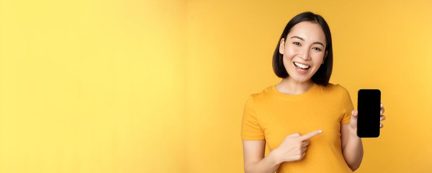 Beautiful happy asian girl showing mobile phone screen, application on smartphone gadget, standing over yellow background.