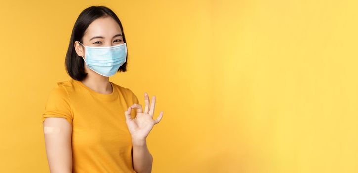 Vaccination from covid and health concept. Happy asian girl showing okay, wearing medical mask, band aid on shoulder, got coronavirus vaccine shot, yellow background.