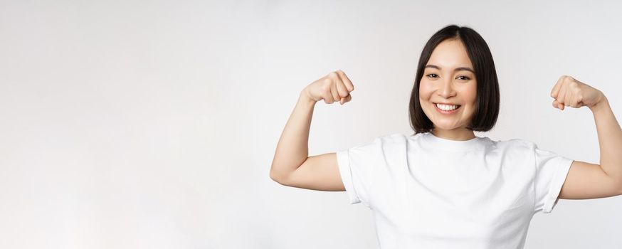 Smiling asian woman showing flexing biceps, muscles strong arms gesture, standing in white tshirt over white background.