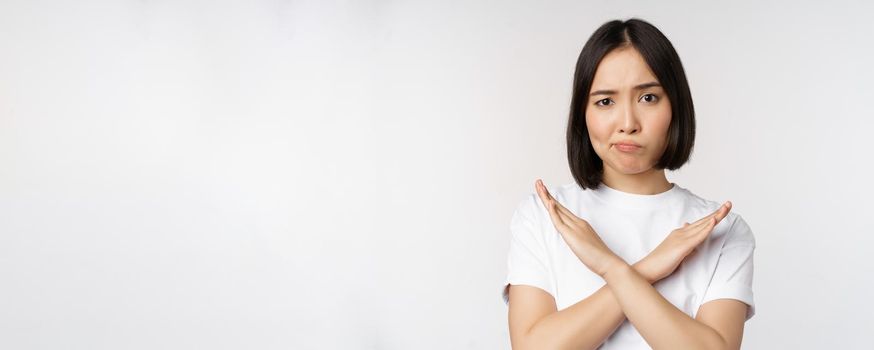 Portrait of asian korean woman showing stop, prohibition gesture, showing arm cross sign, standing in tshirt over white background. Copy space