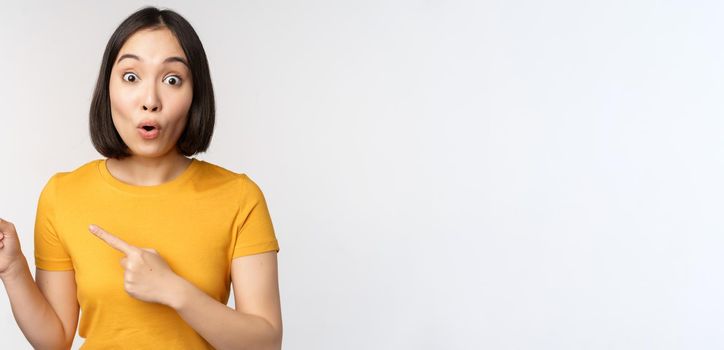 Portrait of smiling asian brunette girl in yellow tshirt, pointing fingers left, showing copy space, promo deal, demonstrating banner, standing over white background.