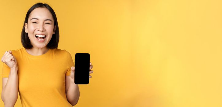 Excited asian woman, showing smartphone app and triumphing, celebrating on mobile phone, standing over yellow background. Copy space