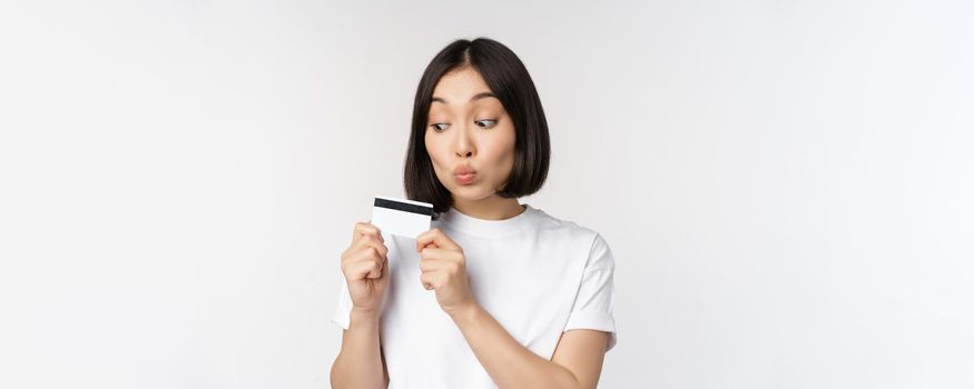 Money and finance concept. Cute japanese girl kissing her credit card, standing in tshirt over white background.