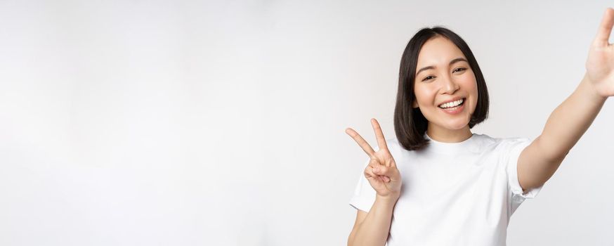 Beautiful young asian woman taking selfie, posing with peace v-sign, smiling happy, take photo, posing against white background.